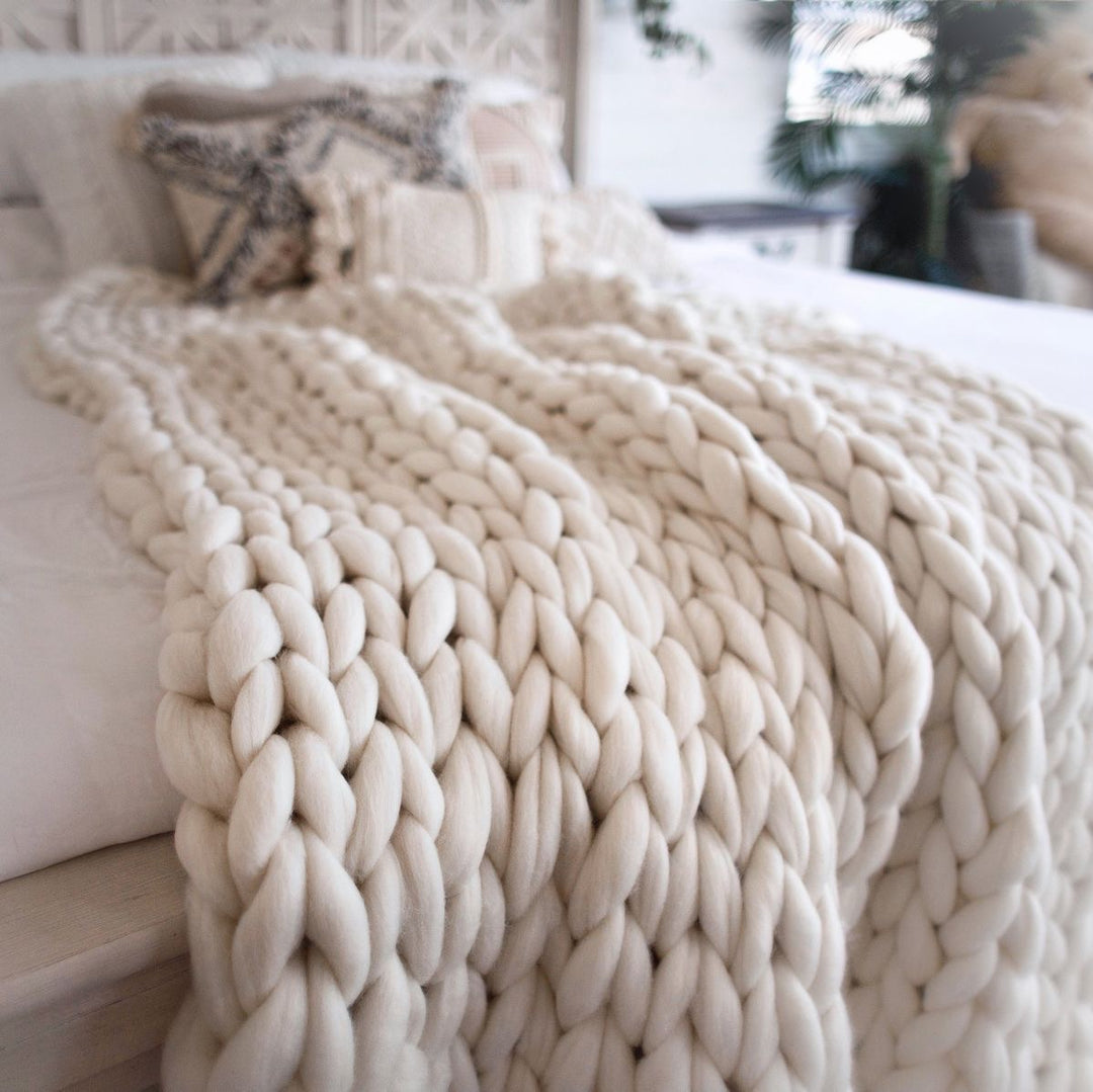 Cozy bed with a large beige knitted blanket and patterned pillow.