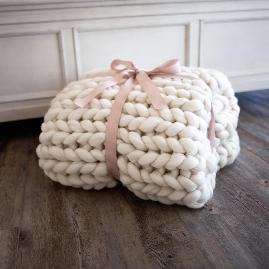 Large cream-colored knitted pouf with a pink ribbon on a wooden floor.
